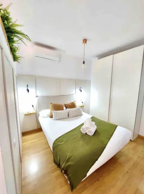 Moderno dormitorio en apartamento de Málaga Centro con cama matrimonial, ropa de cama verde, almohadas blancas, suelo de madera y planta helecho