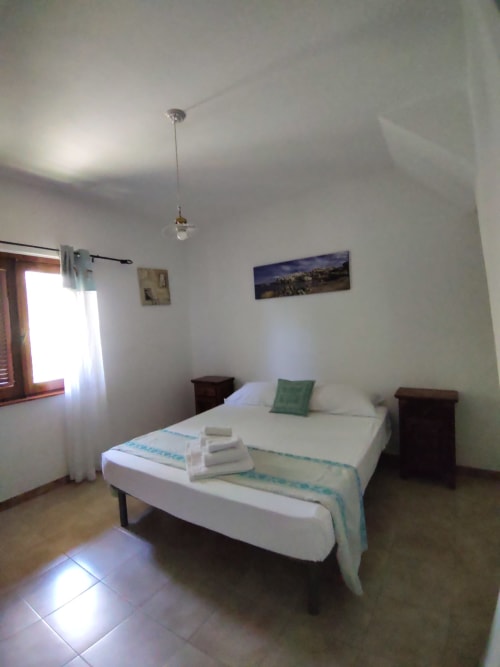 Bright bedroom in San Teodoro villa with queen bed, white linens, green pillow, wooden nightstands, and garden view window