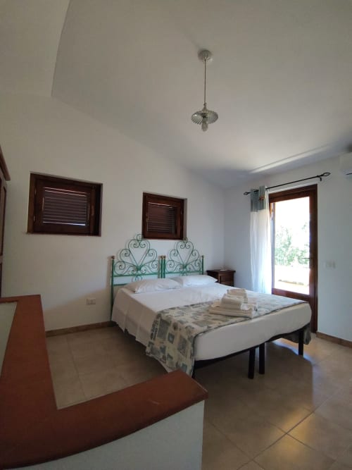Bright bedroom in San Teodoro villa with ornate iron bed frame, white linens, dark wood windows, and glass door opening to