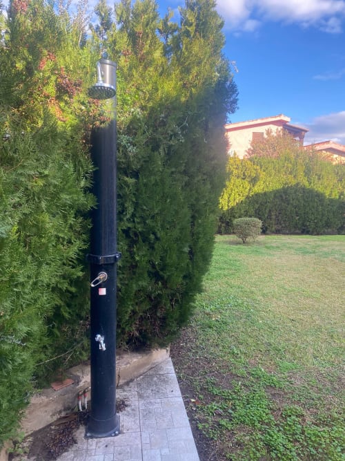 Outdoor shower pole with showerhead in San Teodoro villa garden, surrounded by tall cypress trees and manicured hedges