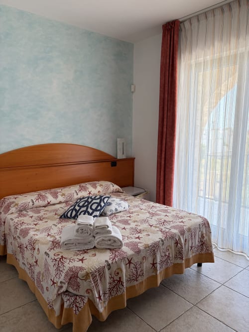 Camera luminosa a Villa Arneide B&B con letto in legno, biancheria a tema marino, tende rosse e vista sul giardino a Torre Lapillo