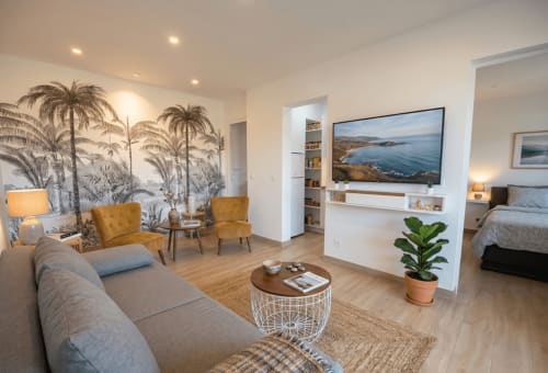 Salon moderne d'appartement avec vue côtière, téléviseur, chaises dorées, fresque palmiers et accès chambre à Conciergerie IB