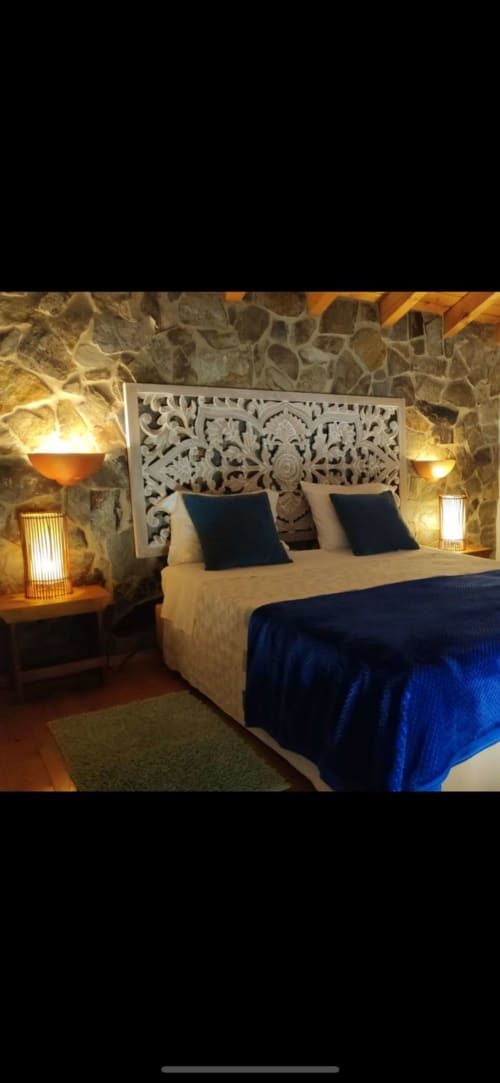 Quarto luxuoso em T Monte Porto Côvo com cabeceira branca ornamentada, almofadas azul marinho, paredes de pedra e iluminação ambiente aconchegante