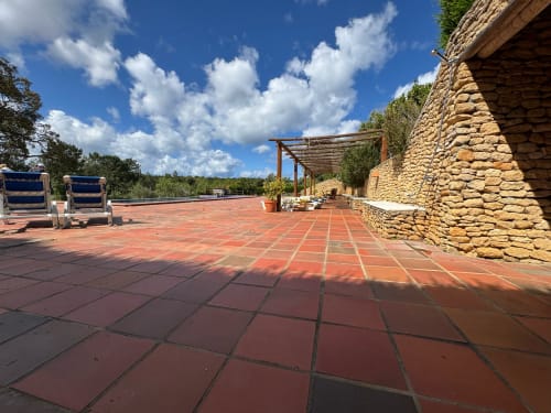 Terraço T Monte em Porto Côvo com pátio de azulejos vermelhos, muros de pedra, espreguiçadeiras azuis e vista para o jardim sob céu azul