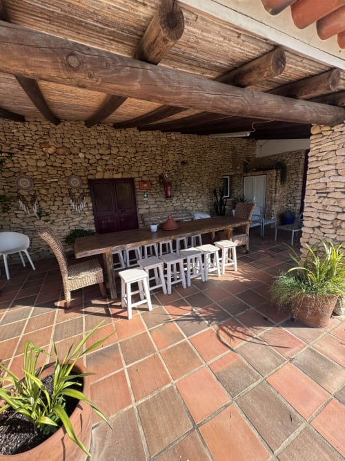 Pátio coberto rústico em T Monte Porto Côvo com vigas de madeira, paredes de pedra, longa mesa de jantar e bancos brancos