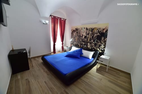 Camera moderna dell'hotel Varigotti Fabio con letto blu, opera d'arte con veduta cittadina, tende rosse e pavimento in legno