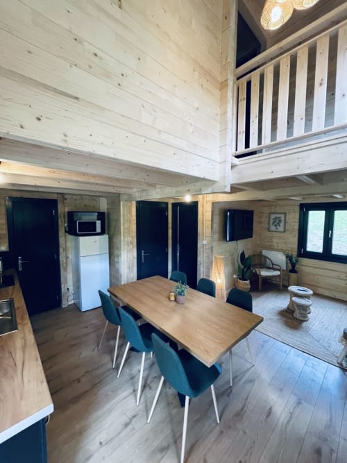 Modern lodge interior with wooden beams, dining table, kitchenette, and upper mezzanine at Cabanes du Belvédère