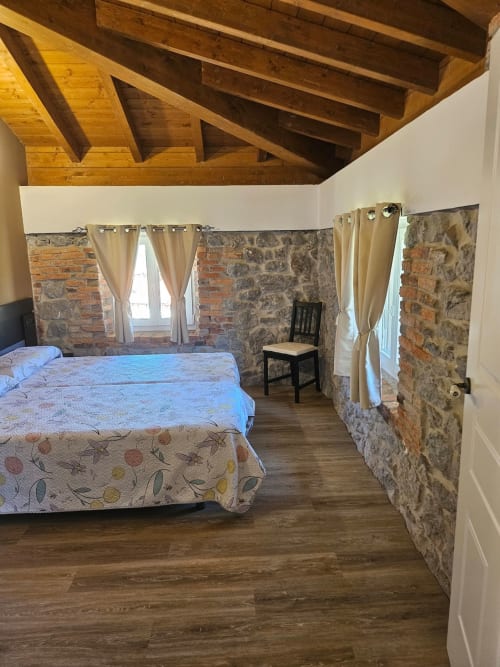 Rustic bedroom at Fabrica de Atardeceres with exposed stone walls, wooden beams, bed, and cream curtains