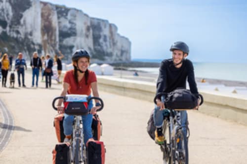 Deux cyclistes pédalent sur la plage près des falaises blanches d'Étretat, France, avec des touristes et la mer en arrière-plan