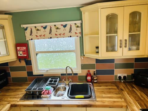 Kitchen sink area at Postwood Gardens Campsite with wooden counters, colorful backsplash, and bird-patterned window blind.