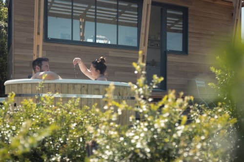 Couple se relaxant dans le bain à remous de Pépite BnB à Villeneuve, avec le lodge moderne en bois et fleurs en arrière-plan