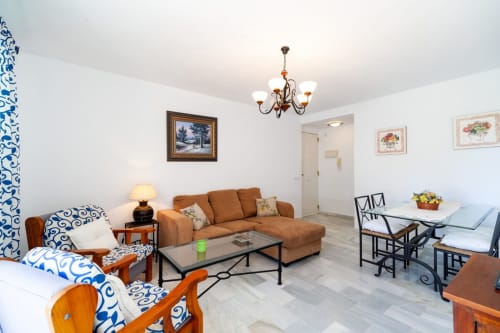 Sala de estar del apartamento Barrosa con sofá marrón, sillas estampadas azules, mesa de comedor y lámpara de araña.