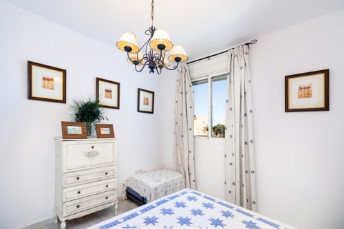 Luminoso dormitorio en apartamento Barrosa Home con ropa de cama estampada azul, cómoda blanca, cuadros enmarcados y balcón con vistas al mar