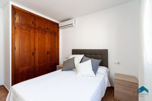 Dormitorio luminoso en Barrosa Home con ropa de cama blanca, cabecero gris, armario de madera y aire acondicionado