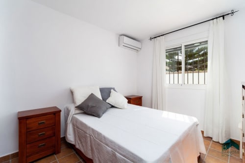 Dormitorio luminoso en villa Barrosa Home con ropa de cama blanca, mesillas de madera, aire acondicionado y puertas de vidrio con vistas al jardín