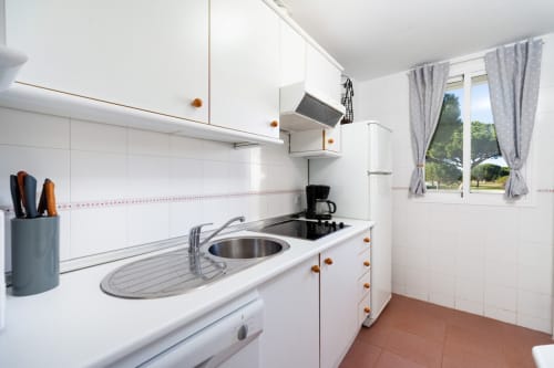 Cocina moderna y compacta en apartamento Barrosa Home con muebles blancos, fregadero de acero inoxidable y piso de baldosas terracota