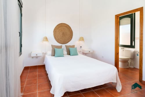 Dormitorio luminoso en Barrosa Home con cama queen, almohadas turquesas, cabecera tejida artesanal, pisos en terracota y baño privado