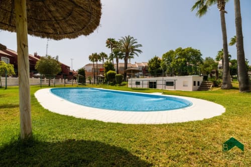 Piscina ovalada con terraza blanca rodeada de césped verde exuberante, palmeras y moderno edificio blanco en Barrosa Home