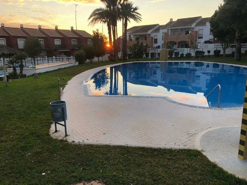 Complejo residencial Barrosa al atardecer con piscina ovalada, palmeras y casas adosadas blancas en España