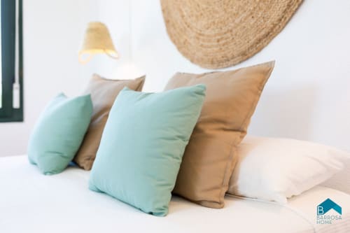 Cómodo dormitorio en Barrosa Home con almohadas turquesas y tostadas sobre ropa blanca, sombrero de paja y lámpara colgante