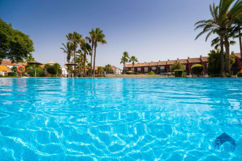 Piscina de aguas cristalinas en el apartamento Barrosa Home en España, rodeada de palmeras y edificios de estilo colonial