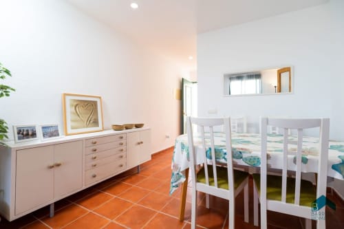 Comedor luminoso del apartamento Barrosa Home con sillas blancas, piso de barro cocido y aparador color crema con decoración