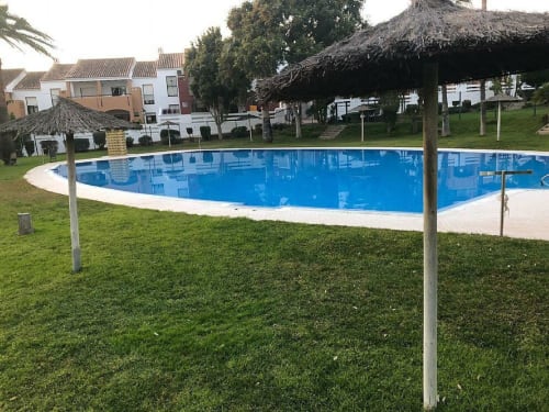Piscina de la comunidad Barrosa Home con agua azul, terraza blanca, estructuras con techo de palma y edificios residenciales