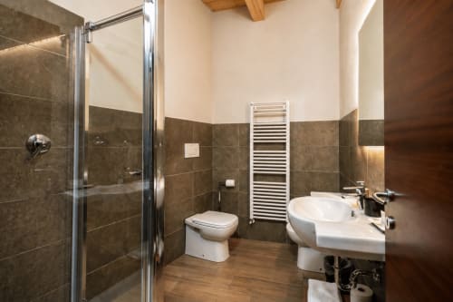 Bagno moderno con rivestimenti in piastrelle scure, lavabo bianco, toilette, doccia a pioggia e scaldasalviette a Al Battistero Luxury