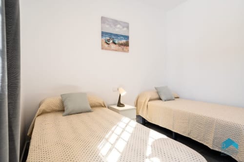 Dormitorio doble en villa Barrosa Home con ropa de cama beige, cuadros de playa y luz natural de las ventanas