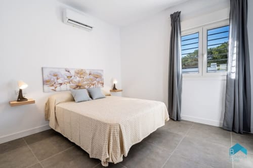 Dormitorio moderno en villa Barrosa Home con cama queen, ropa de cama color crema, cortinas grises y puertas de vidrio con vista al jardín
