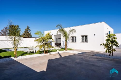 Villa blanca moderna en Barrosa Home con palmeras, jardín cuidado y acceso con patio bajo cielo azul despejado