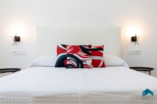 Dormitorio moderno en Barrosa Home con cabecera acolchada blanca, almohada decorativa roja y negra, y apliques laterales.