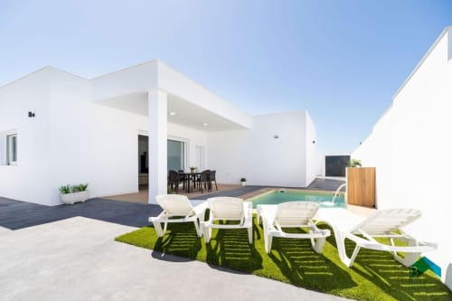 Villa Bison A en Barrosa Home presenta arquitectura blanca moderna con tumbonas, pequeña piscina, zona de comedor y jardines cuidados