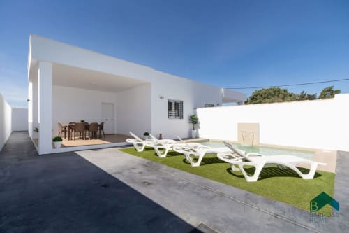 Villa moderna blanca con patio cubierto, comedor y tumbonas junto a la piscina en Barrosa Home, España
