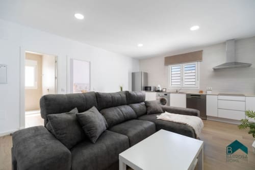 Apartamento moderno diáfano en Barrosa Home con sofá gris carbón, mesa de café blanca y cocina integrada en acero inoxidable