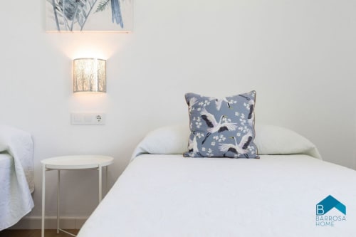 Dormitorio moderno en Barrosa Home con cama blanca, almohada azul con estampado de pájaros, mesilla y aplique de pared