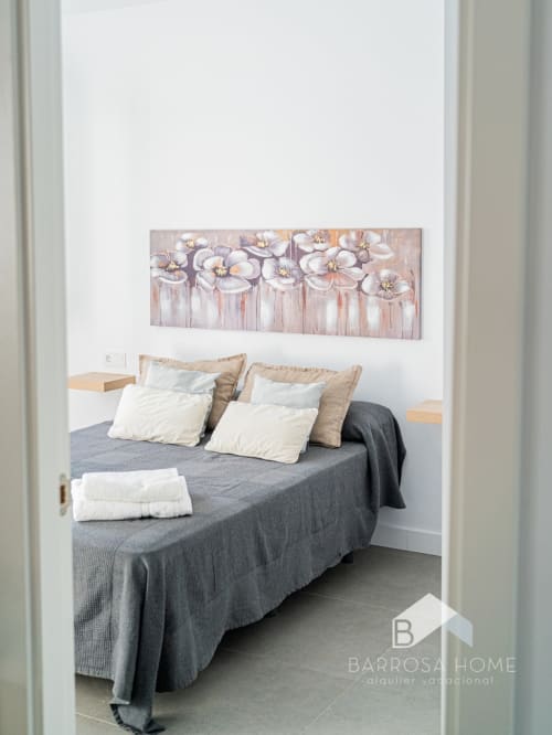Dormitorio moderno en villa Barrosa Home con cama gris, almohadas blancas y obras de arte floral en paredes blancas
