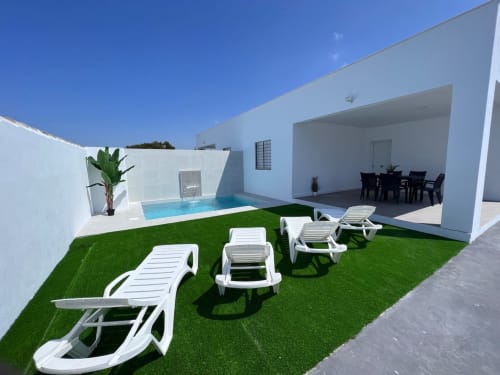 Villa moderna con piscina, tumbonas blancas sobre césped artificial y terraza cubierta con zona de comedor en Barrosa Home