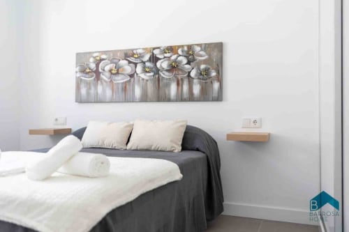 Dormitorio moderno en villa Barrosa Home con cuadro de flores blancas, cama oscura, mesitas de madera y decoración neutral