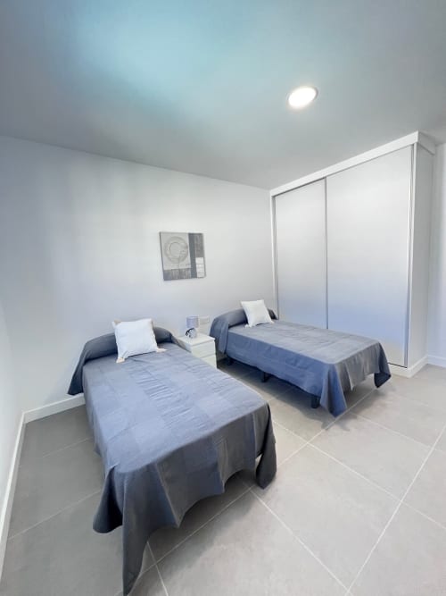 Moderno dormitorio doble en villa Barrosa Home con camas azules, almohadas blancas, armario corredizo y piso de baldosas