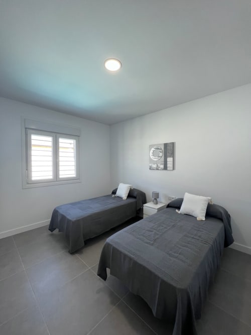 Dormitorio doble en Villa Granizo de Barrosa Home con camas oscuras, almohadas blancas, ventana con persianas e iluminación empotrada
