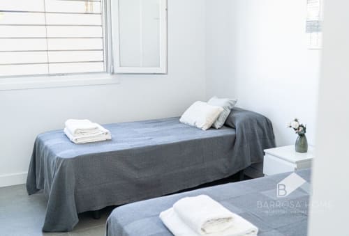 Dormitorio moderno en Barrosa Home con camas gemelas, ropa de cama gris, almohadas blancas, mesita de noche con flores y ventanas luminosas