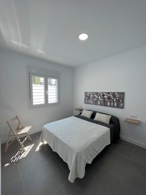 Dormitorio moderno en villa Barrosa Home con cama matrimonial, ventana con contraventanas, silla de madera y piso de azulejos oscuros