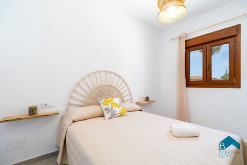 Dormitorio luminoso en villa Barrosa Home con cabecero tejido, ropa de cama crema, ventanas de madera y luz natural desde el techo