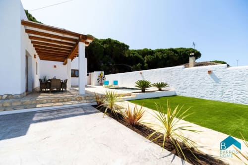 Villa Barrosa con paredes blancas, pérgola de madera, piscina, césped verde y paisajismo mediterráneo