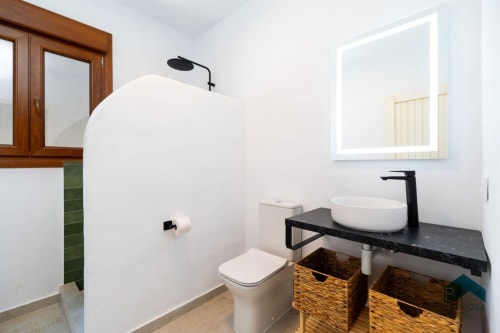 Baño moderno de villa Barrosa Home con lavabo blanco, grifo negro, canastas tejidas y espejo iluminado