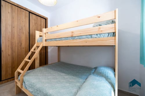Litera de madera clara con ropa de cama azul en apartamento Barrosa Home, con armario de madera y luminaria moderna de techo