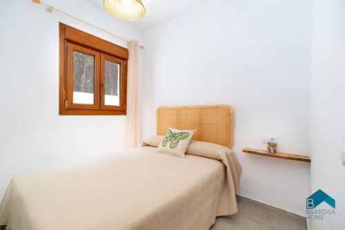 Luminoso dormitorio en Barrosa Home con cama de cabecero de madera, silla de ratán y ventana con vistas al jardín