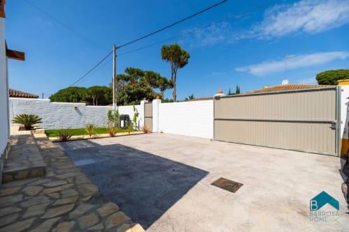 Entrada de villa Barrosa con muros blancos, portón metálico, estacionamiento y jardín mediterráneo bajo cielo azul