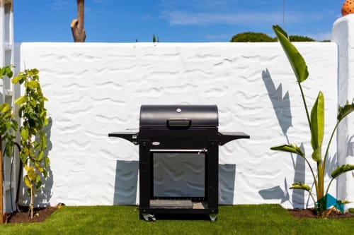 Parrilla negra de barbacoa sobre el césped frente a pared texturizada blanca en villa Barrosa Home con plantas tropicales